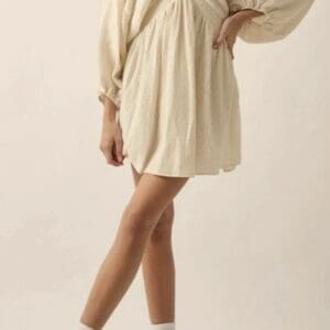 Person wearing a cream-colored, long-sleeve, V-neck mini dress with white socks and light-colored high-top sneakers, standing with one leg crossed over the other.