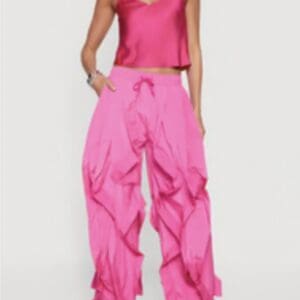 A woman wears a bright pink satin camisole top and loose, ruffled pink pants with drawstrings, paired with strappy heels.