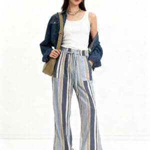 Woman standing against a white background, wearing a white tank top, striped wide-leg pants, denim jacket, white cap, sneakers, necklaces, and a crossbody bag.