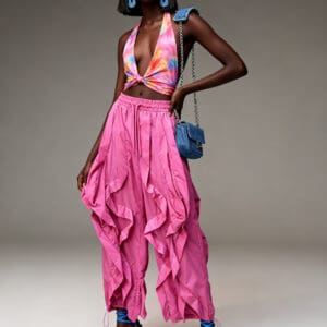 Woman posing in a colorful halter top, pink ruffled pants, blue high heels, large blue earrings, and holding a small blue purse against a plain background.