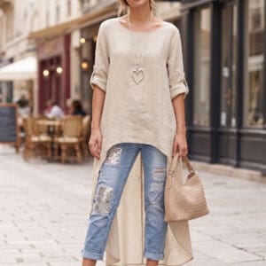 Woman standing on a cobblestone street wearing a beige long tunic, distressed jeans with metallic patches, nude heeled sandals, and holding a woven handbag. Outdoor café seating in the background.