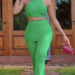 Person wearing a bright green sleeveless crop top and matching high-waisted pants, holding a small pink handbag, standing outdoors in front of wooden doors.