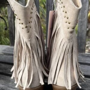 A pair of beige suede cowboy boots with fringe, gold studs, and stacked wooden heels displayed on a wooden surface.