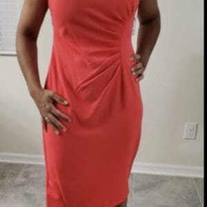 Woman wearing a sleeveless, knee-length red dress with side ruching, standing indoors on a tiled floor.