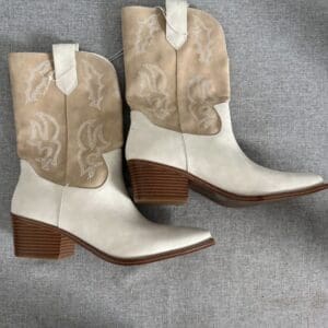 A pair of two-tone, beige and cream cowboy boots with stitched detailing and medium stacked wooden heels placed on a grey fabric surface.