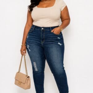 Woman wearing a beige top, high-waisted blue jeans, wedge sandals, and carrying a beige quilted handbag, standing against a white background.