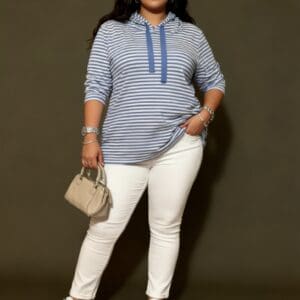 Woman wearing a blue and white striped hoodie, white pants, and blue sneakers, holding a beige handbag, standing against a plain background.