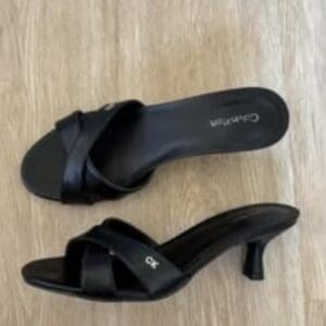 A pair of black, open-toe heeled sandals with crisscross straps are placed on a wooden floor.