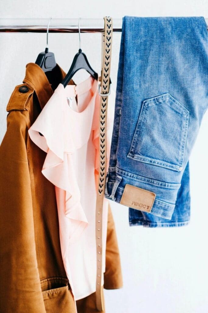 A brown coat, a pale pink ruffled blouse, a patterned beige belt, and a pair of blue jeans are hanging on a clothing rack against a white background.