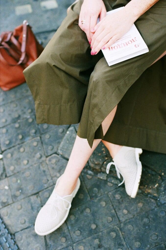 Person wearing olive green skirt and white shoes sitting on a bench, holding a book with a brown bag on the ground nearby.