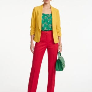 A woman wearing a yellow blazer, a green floral top, and red trousers, holding a green handbag.