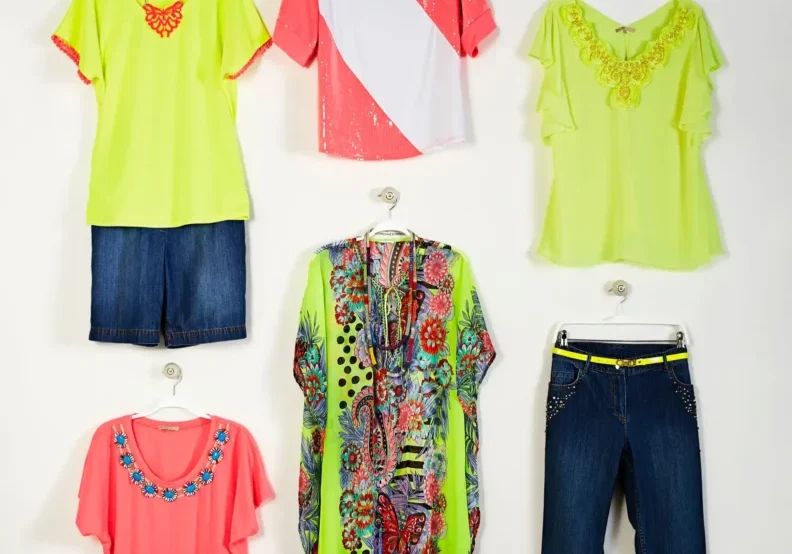Six colorful women's outfits including tops, shorts, a kaftan, and jeans are displayed on hangers against a plain white background.