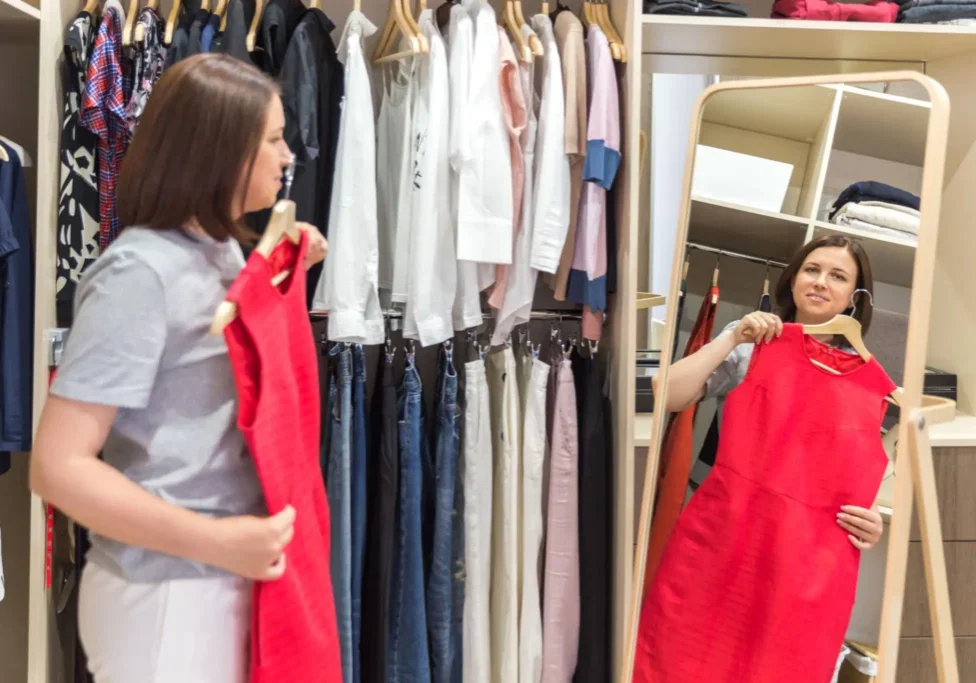 A woman stands in front of a mirror in a walk-in closet, holding a red dress up to herself as she considers it.