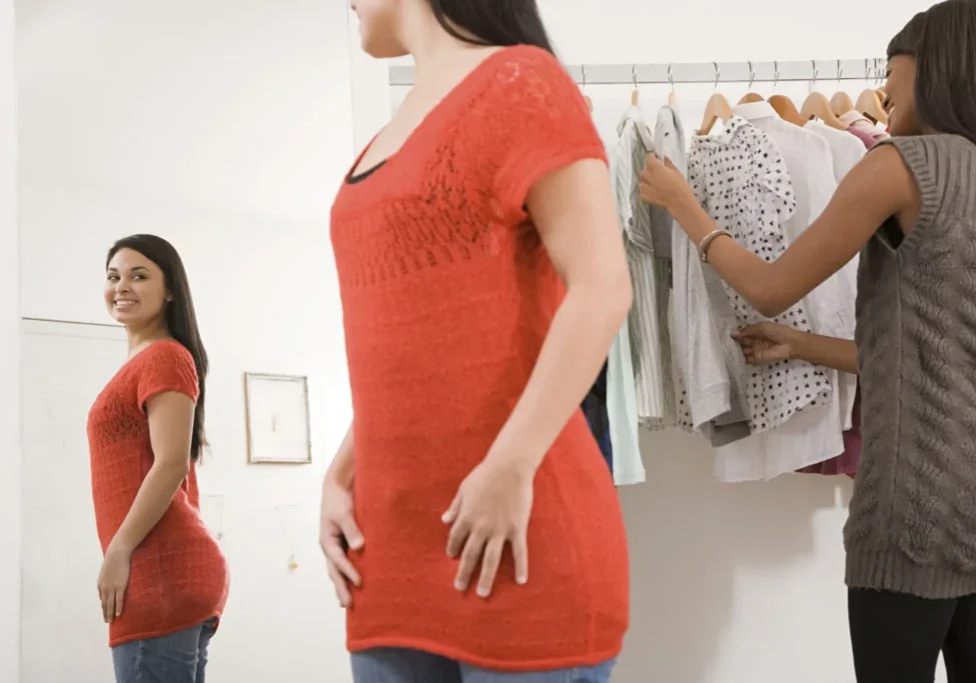A woman in a red top looks at her reflection in a mirror while another woman browses clothes on a rack in a store.
