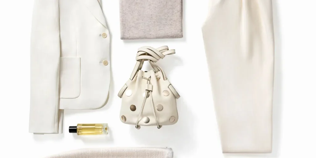 A flat lay of cream and beige clothing and accessories, including a blazer, turtleneck, trousers, skirt, purse, shoes, sunglasses, earrings, and a perfume bottle.