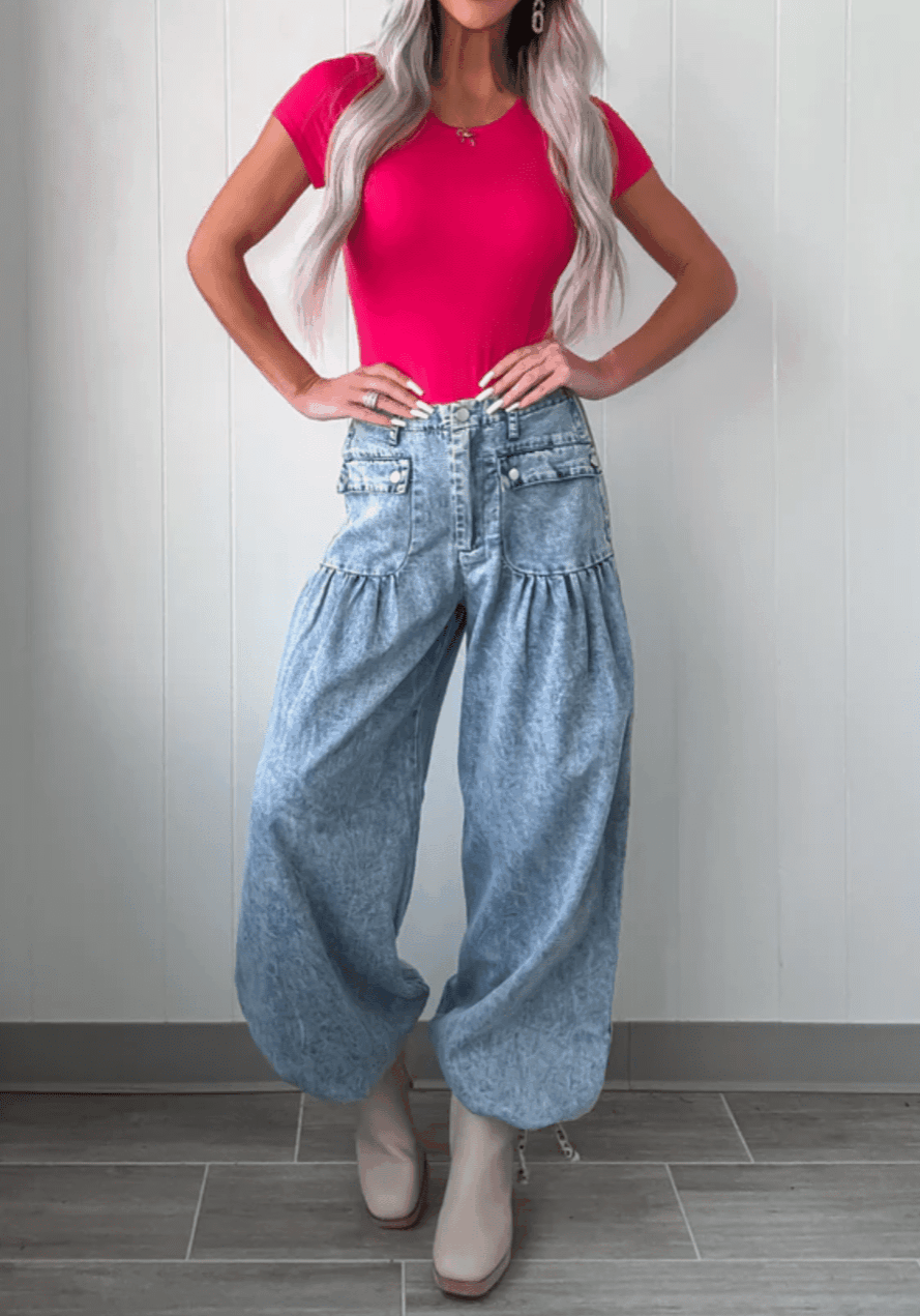 A person stands against a light-colored wall wearing a bright pink fitted short-sleeve top, 6 Button Pocket Dress Pants in a high-waisted baggy light-wash style, and beige platform boots.