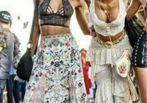 Two women at an outdoor festival wear patterned skirts and crop tops, posing and smiling for the photo. Other festival attendees and a stage are seen in the background.