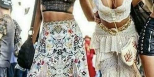 Two women at an outdoor festival wear patterned skirts and crop tops, posing and smiling for the photo. Other festival attendees and a stage are seen in the background.