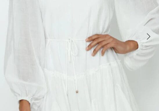 A woman wearing a long-sleeved, white, ruffled mini dress with a cinched waist, standing against a plain light background.