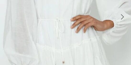 A woman wearing a long-sleeved, white, ruffled mini dress with a cinched waist, standing against a plain light background.