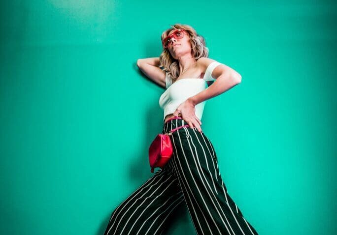 A woman poses against a teal wall wearing striped wide-leg pants and a white top, with a neon sign reading "good vibes only" glowing above her.