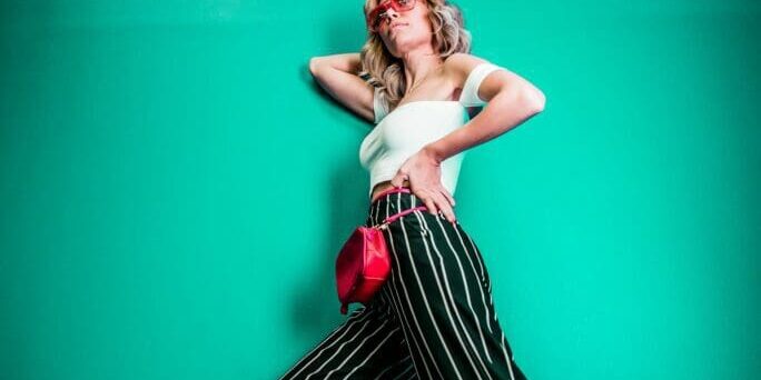 A woman poses against a teal wall wearing striped wide-leg pants and a white top, with a neon sign reading "good vibes only" glowing above her.