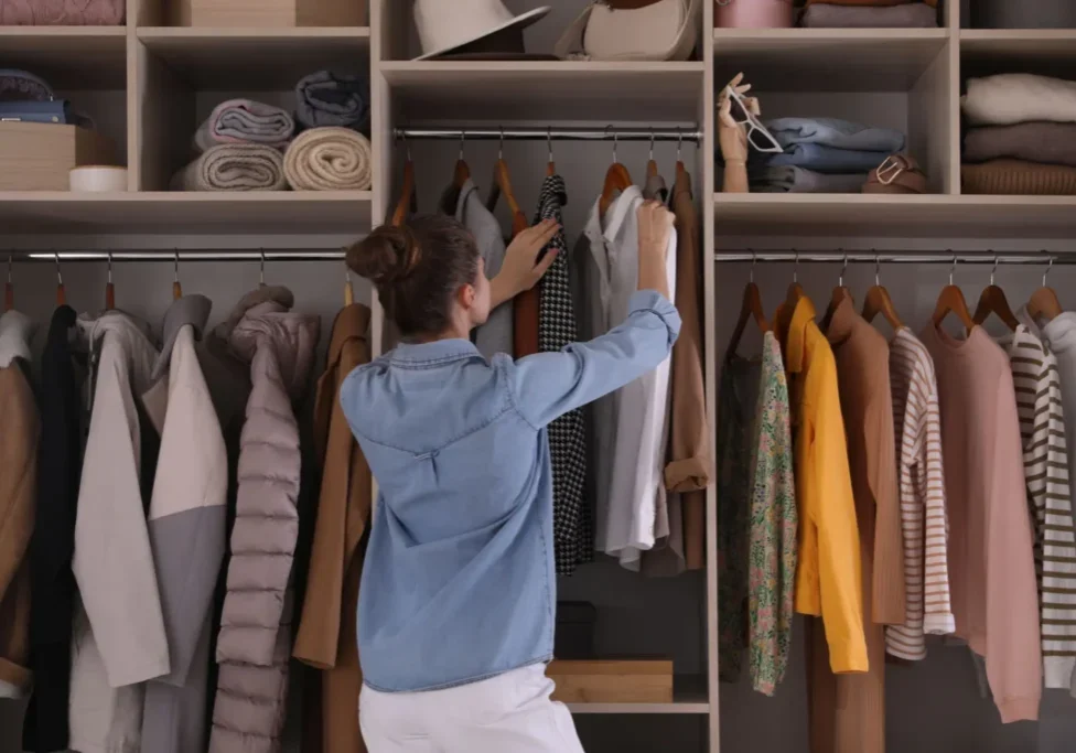 A woman stands in front of an organized closet, reaching for a shirt among neatly hung clothes and folded items on shelves.