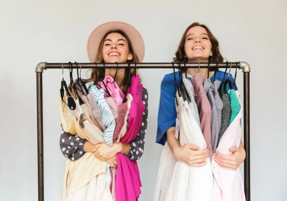 Two women stand behind a clothing rack, smiling and holding various colorful garments draped over hangers.