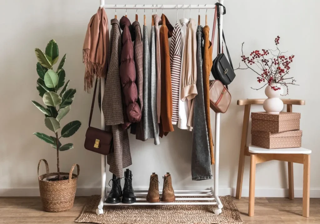 A white clothing rack holds various jackets, tops, and bags. Boots sit below. Nearby are a potted plant, stacked boxes on a chair, and a vase with red berries.