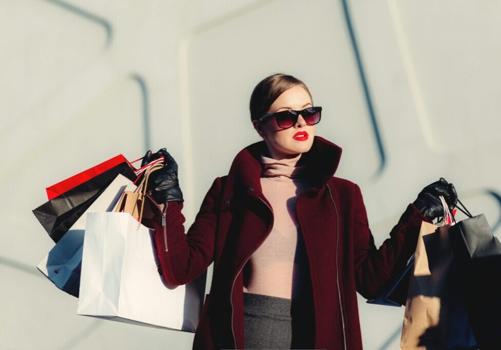 A woman wearing sunglasses and a dark coat holds multiple shopping bags in both hands, standing against a light-colored, modern wall.