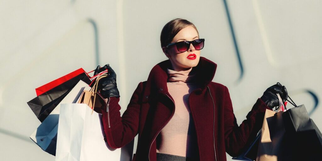 A woman wearing sunglasses and a dark coat holds multiple shopping bags in both hands, standing against a light-colored, modern wall.