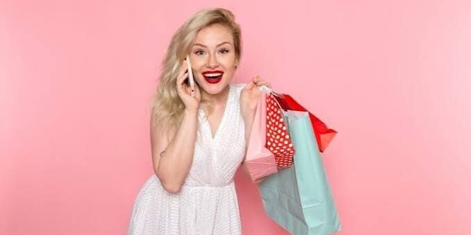 A woman in a white dress smiles while holding shopping bags and talking on a phone against a pink background.