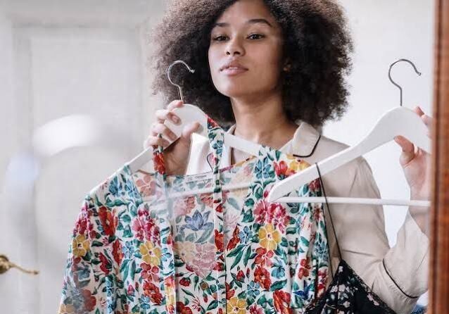 A person stands in front of a mirror holding up two floral print dresses on hangers, appearing to decide which one to wear.