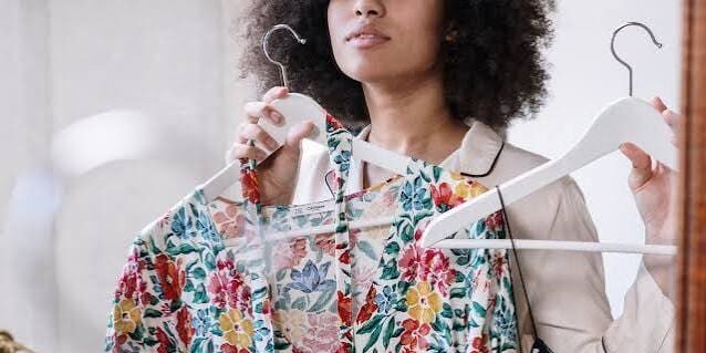A person stands in front of a mirror holding up two floral print dresses on hangers, appearing to decide which one to wear.
