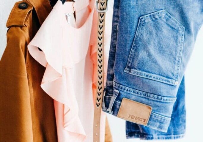 A brown coat, a pale pink ruffled blouse, a patterned beige belt, and a pair of blue jeans are hanging on a clothing rack against a white background.