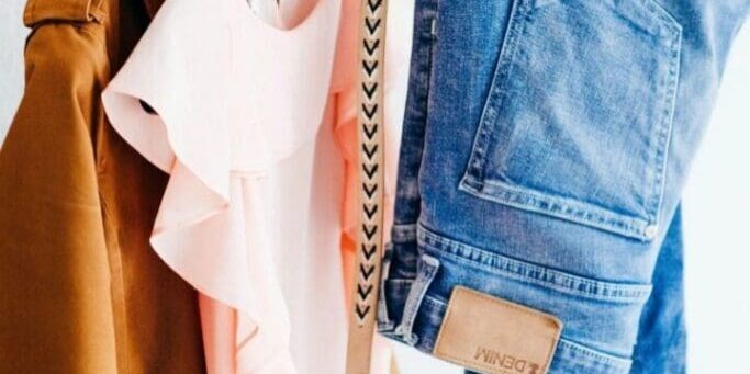 A brown coat, a pale pink ruffled blouse, a patterned beige belt, and a pair of blue jeans are hanging on a clothing rack against a white background.