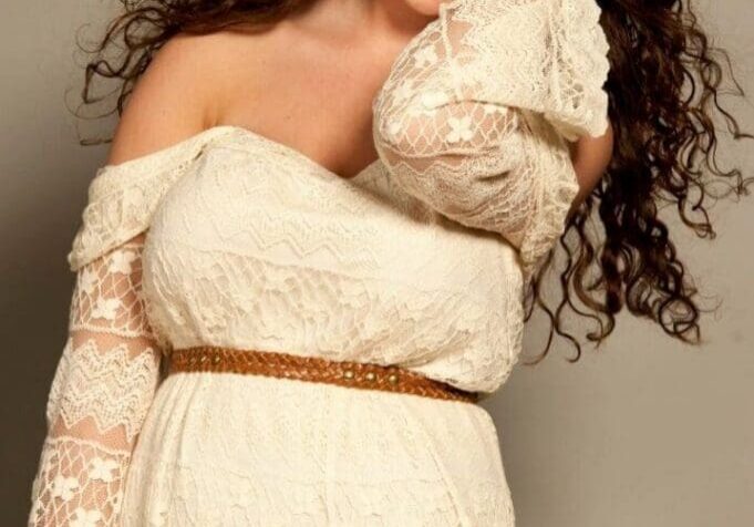Woman with long curly hair wearing an off-shoulder white lace dress and brown belt, smiling and touching her hair against a neutral background.