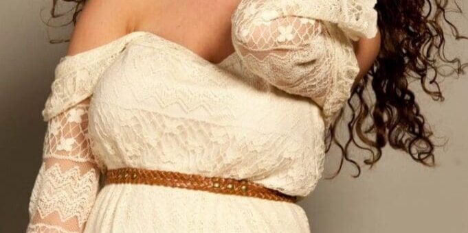Woman with long curly hair wearing an off-shoulder white lace dress and brown belt, smiling and touching her hair against a neutral background.