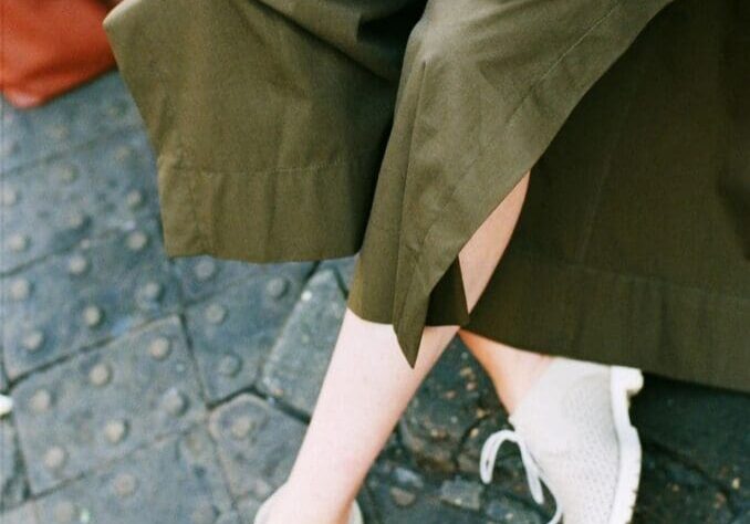 Person wearing olive green skirt and white shoes sitting on a bench, holding a book with a brown bag on the ground nearby.