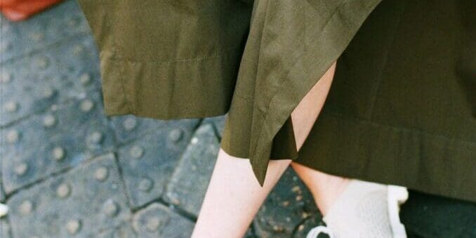 Person wearing olive green skirt and white shoes sitting on a bench, holding a book with a brown bag on the ground nearby.