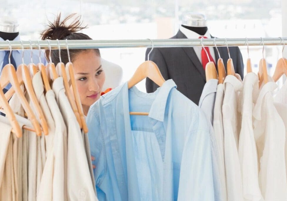 A woman looks through a rack of light-colored shirts on hangers in a clothing store, with mannequins dressed in suits in the background.