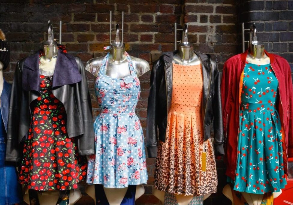 Four mannequins display colorful dresses and jackets against a brick wall, each featuring different patterns, including floral and polka dots.
