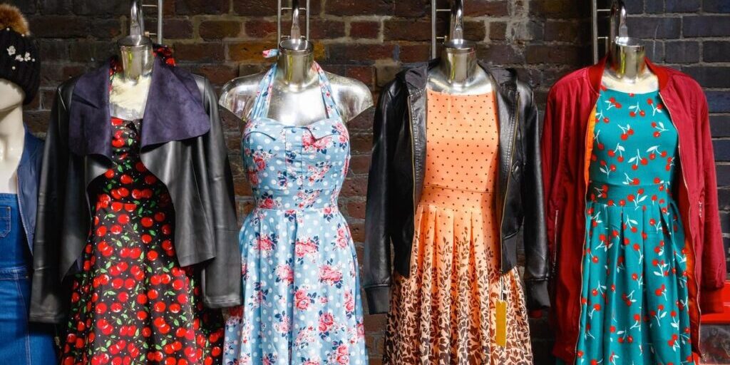 Four mannequins display colorful dresses and jackets against a brick wall, each featuring different patterns, including floral and polka dots.