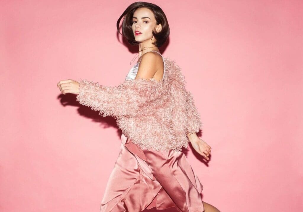 A woman in a pink fuzzy jacket and satin skirt walks in front of a solid pink background.