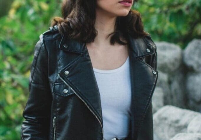 A woman with shoulder-length dark hair wearing a black leather jacket, white top, and blue jeans stands outdoors near rocks and greenery.