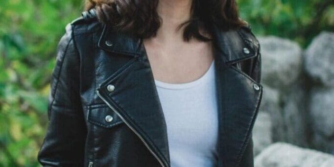 A woman with shoulder-length dark hair wearing a black leather jacket, white top, and blue jeans stands outdoors near rocks and greenery.