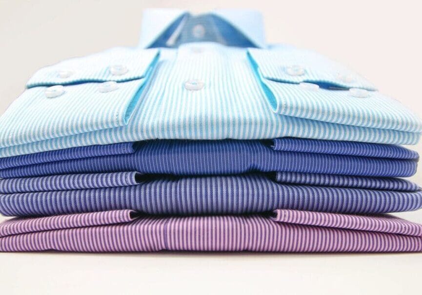 Five neatly folded dress shirts in shades of blue and purple, stacked on top of each other, viewed from the front.