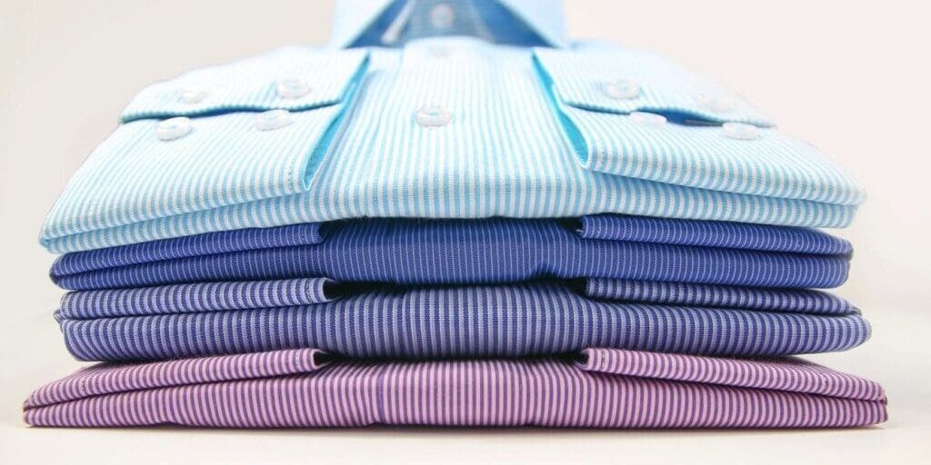 Five neatly folded dress shirts in shades of blue and purple, stacked on top of each other, viewed from the front.