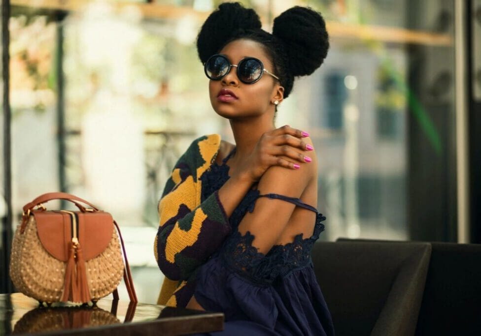 Woman with natural hair in two puffs wearing sunglasses, a dark blue dress, and a patterned sweater sits at a table with a woven handbag beside her.