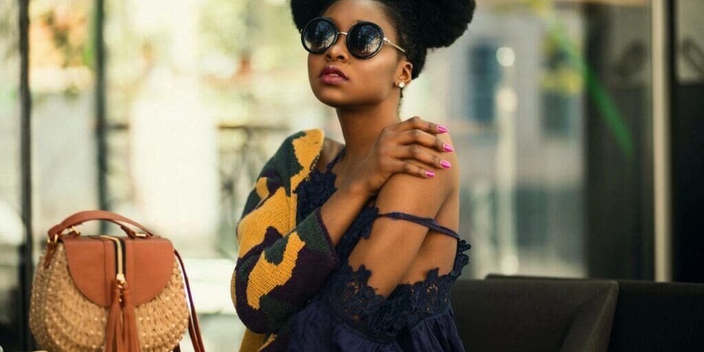 Woman with natural hair in two puffs wearing sunglasses, a dark blue dress, and a patterned sweater sits at a table with a woven handbag beside her.